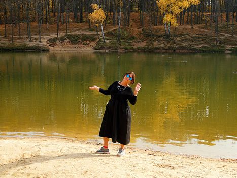 smiling woman in sunglasses and black loose dress posing on the shore of an autumn lake