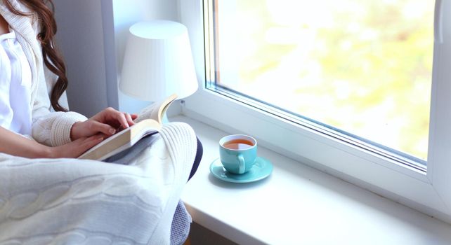 Young woman at home sitting near window relaxing in her living room reading book and drinking coffee or tea . Young woman at home .