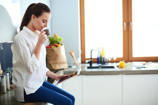 Young woman using tablet in kitchen at home and drinking coffee.