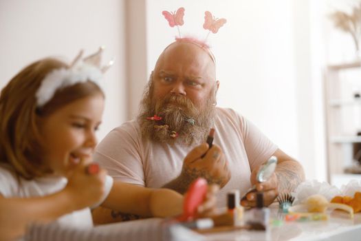 Emotional bearded man with butterflies on headband looks at adorable little girl applying colorful lipstick at table in living room. Beauty salon at home