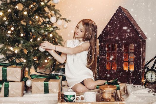 Little girl in white dress decorating Christmas tree with white balls under snowfall.