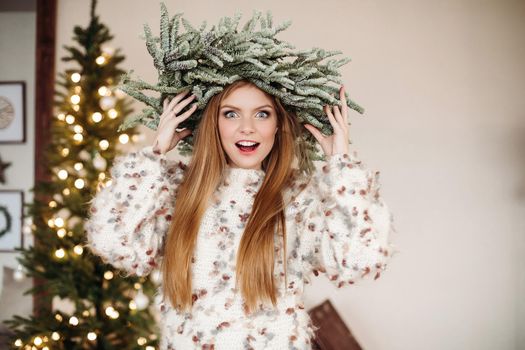 Pretty redhead wearing beautiful Christmas wreath on head and looking at camera with surprise.