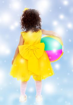 Very small curly-haired girl in bright yellow dress with a huge bow , turned her back to the camera. Girl holding under one arm the ball.Blue winter background with white snowflakes.