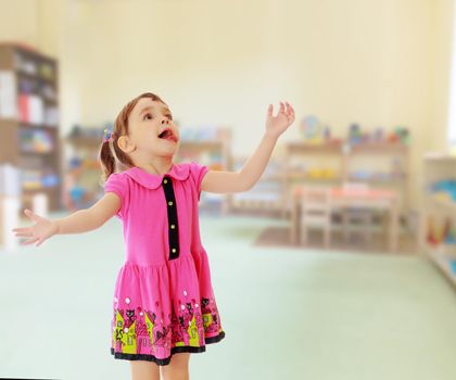 Surprised little girl raised his head up and gesturing with his hands. Close-up.On blurred background the great hall of the kindergarten, with long racks where there are toys.