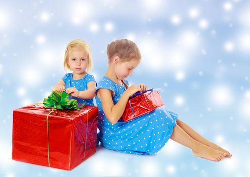 Two charming little girls , sisters, in identical blue dresses with polka dots. Girl looking at gifts Packed in beautiful red paper tied with a bow.On a blue background with large, white, Christmas