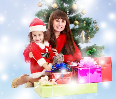 Happy mother and daughter near a Christmas tree surrounded by heaps of gifts.Blue winter background with white snowflakes.