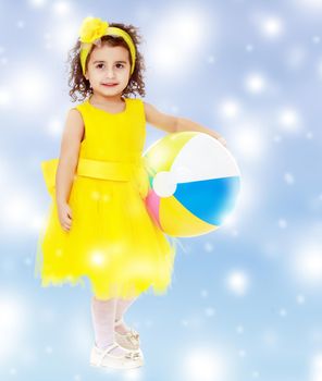 Cute curly girl in a bright yellow dress and bow on her head holding the ball. Close-up.Blue winter background with white snowflakes.