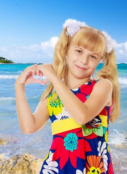 Beautiful little girl with long blond hair braided in ponytails , folded palms heart.On the background of sea beach, warm sea and blue sky with clouds.