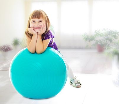 The concept of child rearing in the family and kindergarten. On the background of the hall with large , bright Windows. About what dreams a little girl , put his hands on big blue fitness ball.