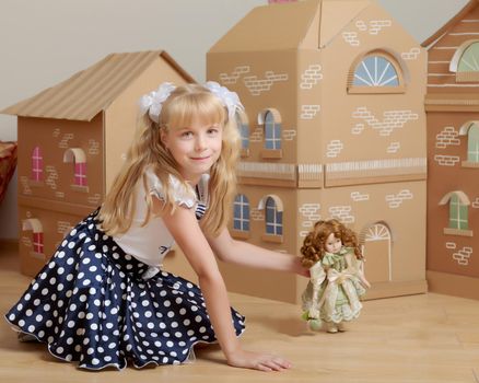 Beautiful little girl with long blonde ponytails on her head , playing with a doll near a cardboard house