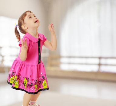 Pensive little girl with pigtails on the head , in a pink dress. The girl was looking at the top turned sideways to the camera.On the background of the great hall of the kindergarten