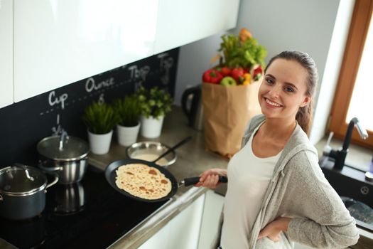 Young woman cooking pancakes at kitchen standing near stove.