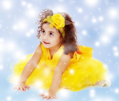 Funny curly little girl in a bright yellow elegant dress crawling on the floor. She looks to the top.Blue winter background with white snowflakes.