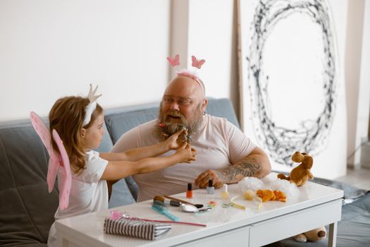 Happy plump father plays beauty salon with little girl wearing fairy suit putting scrunchie onto his beard on sofa at home