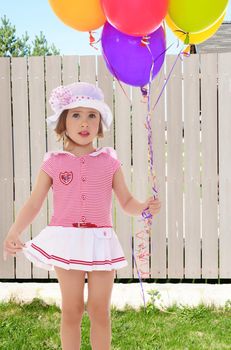 Adorable little girl holding colorful balloons. A girl in a short pink dress and white hat with pink bow.Closeup.On the background of beautiful wooden fence.