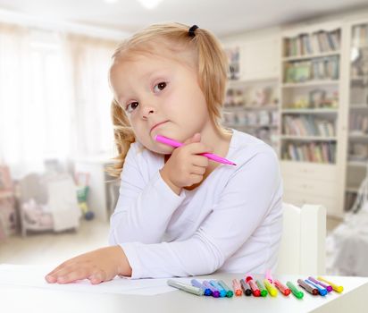 Adorable little blonde girl with white t-shirts without a pattern, draws markers at the table on a white sheet of paper. The girl rested her head on the arm , it's about something thought . close-up