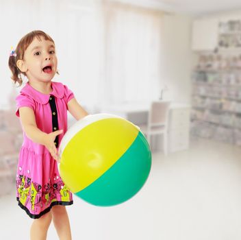 Emotional little girl with pigtails on the head , in a pink dress. Girl catches with hands a large, inflatable striped ball.In the background children's room, where the shelves are containers
