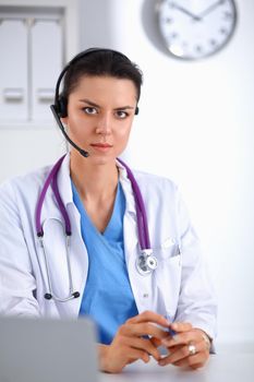 Doctor wearing headset sitting behind a desk with laptop over grey background.