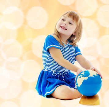 Nice little girl in a Sea blue dress sitting on the floor. Girl turns hand the globe.On a brown background with white blurred circles.