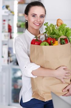 Young woman holding grocery shopping bag with vegetables .Standing in the kitchen. Woman in the kitchen looking at the camera.