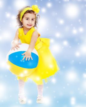 Funny curly baby girl in a bright yellow dress and bow on her head playing with a ball.Blue winter background with white snowflakes.