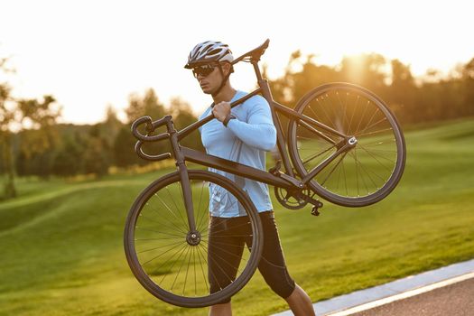 Professional road bicycle racer. Strong athletic man in sportswear and protective helmet carrying his bike after after training in park at sunset. Active lifestyle and sport concept. Biking outdoors