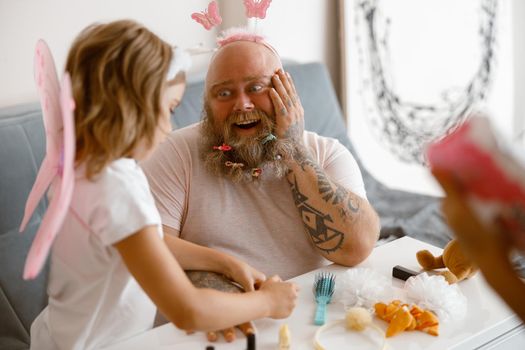 Surprised bearded father with headband and scrunchies looks at little daughter in fairy suit at table with different accessories in living room. Playing beauty salon