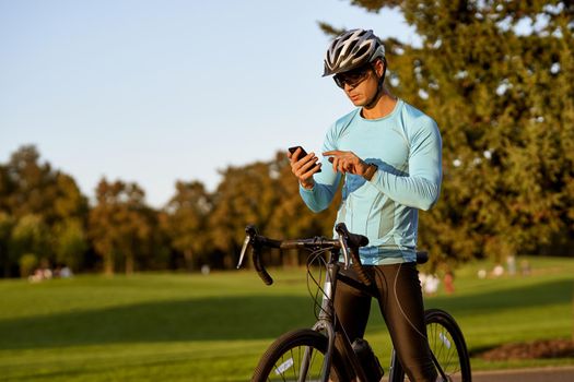 Checking results. Young athletic man, professional cyclist in sportswear and protective helmet standing with his bike in park and using his smartphone. Active lifestyle and sport