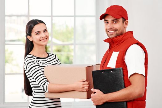 Cheerful postman wearing red postal uniform is delivering parcel to a satisfied client. He holds delivery report after receing a sign from the client. Dark-haired woman holds carton box and looking at the camera with a smile. Friendly worker, high quality delivery service. Indoors. Bright interior.