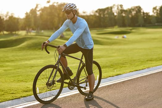 Summer cycling training. Young athletic man in sportswear riding bicycle along a road in city park at sunset, full length. Healthy active lifestyle and sport concept