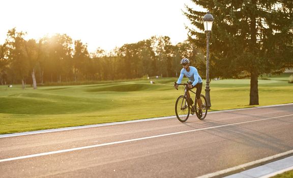 Athletic man in sportswear cycling in city park at sunset, riding bicycle along a road. Healthy active lifestyle and sport concept