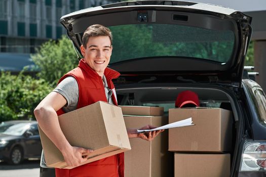 Smiling delivery man in red postal uniform standing near trunk of his car with a box and a clipboard in his hands. He is ready to do his job. Friendly worker, high quality delivery service. Outdoors.