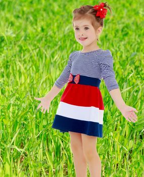 Little Caucasian blond girl with a red bow on her head, dressed in striped dress, she shrugs. Close-up.On blurred background of green grass, concept of summer holidays for children.