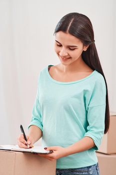 Young entrepreneur, SME, freelance woman working online, business by using smartphone, making purchase, order and preparing packages. Woman standing near stack of boxes, making notes on a clipboard. Concept of success