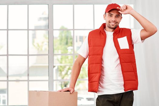 Cheerful postman wearing red postal uniform is delivering parcel to a client. He has done his job very well. He touches his cap and looking at the camera with a smile. Friendly worker, high quality delivery service. Indoors. Bright interior.