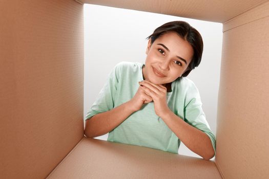 Happy girl unpacking, opening carton box and looking inside. She clasped her hands under her chin. The package, delivery, surprise, gift lifestyle concept. Human emotions and facial expressions concept