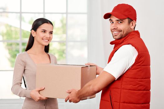 Cheerful postman wearing red postal uniform is delivering parcel to a satisfied client. He is looking at the camera, while she is looking at him. They both hold carton box and smiling. Friendly worker, high quality delivery service. Indoors. Bright interior.