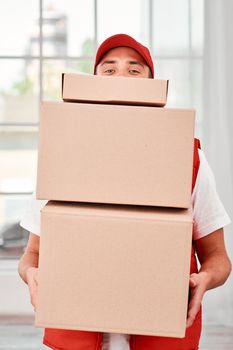 Cheerful postman wearing red postal uniform is delivering parcels to a client. He holds carton boxes and looking at the camera. Friendly worker, high quality delivery service. Indoors. Bright interior.