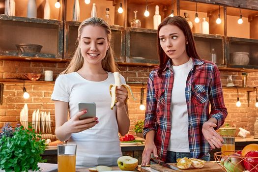 Portrait of two attractive women, having fun, while preparing fruit salad. londe girl in white T-shirt is using her phone while eating a banana. Her disgruntled friend in checkered shirt looks at her and waits when they'll start cooking. Front view