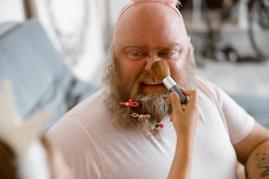 Plump man with scrunchies in beard grimaces while girl applies makeup onto his nose playing beauty salon in contemporary living room