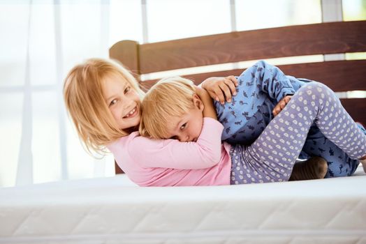 Happy little sister and brother in pajamas playing and having fun while lying on a large comfortable white mattress in bedroom. Happy family at home