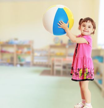 Happy little girl with pigtails on the head , in a pink dress. The girl lifted a large, inflatable striped ball.On blurred background the great hall of the kindergarten