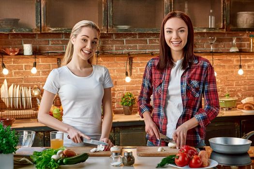 Portrait of two attractive women, having fun, while preparing vegetable salad. They are fully involved in the process. They are looking at the camera and smiling while slicing mushrooms. Front view