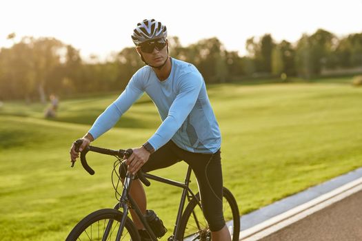 Strong athletic man in sportswear and protective helmet standing with his bicycle on the road in park and looking away, resting after cycling outdoors. Healthy active lifestyle and sport concept