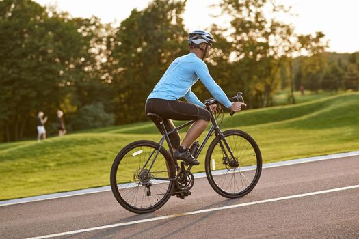 Biking outdoors. Side view of athletic man in sportswear riding bicycle along a road in city park at sunset, full length. Healthy active lifestyle and sport concept