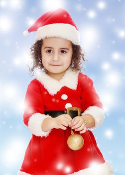Very cute ,with curly hair little girl in a suit and cap of Santa Claus , is holding a Christmas toy. Close-up.Blue winter background with white snowflakes.