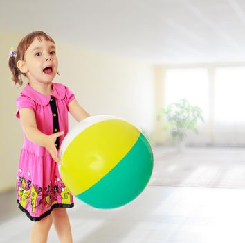 Emotional little girl with pigtails on the head , in a pink dress. Girl catches with hands a large, inflatable striped ball.Against the background of a large room with Windows and flowers .