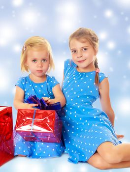 Two charming little girls , sisters, in identical blue dresses with polka dots. Girl looking at gifts Packed in beautiful red paper tied with a bow.On a blue background with large, white, Christmas