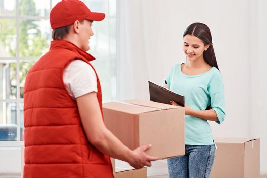 Cheerful man wearing red postal uniform is delivering parcel to a satisfied client. He holds carton box while waiting. Dark-haired woman is signing receipt of delivery package. Friendly worker, high quality delivery service. Indoors.