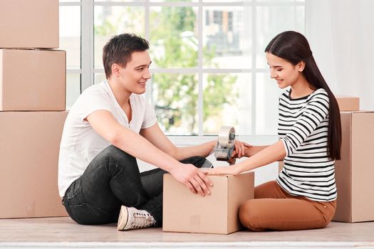 Young couple of freelancers, entrepreneurs, sellers prepare product, parcel box for delivering to customer. They use tape for wrapping parcels. Indoors, bright interior. Online selling, e-commerce concept